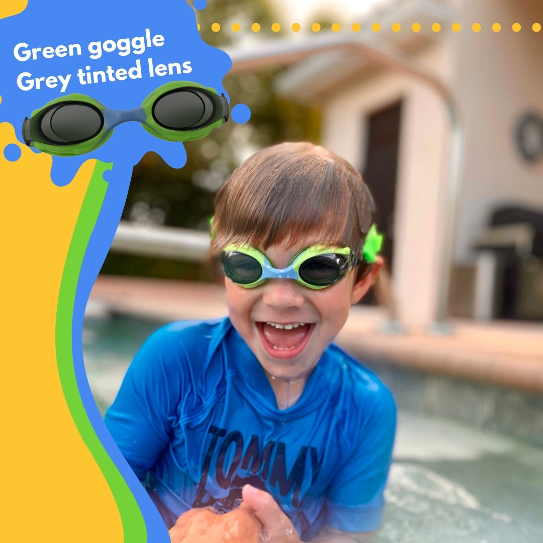 Young boy with blonde hair wearing blue Frogglez swim goggles sitting in swimming pool laughing. Text reads green goggle grey tinted lens.